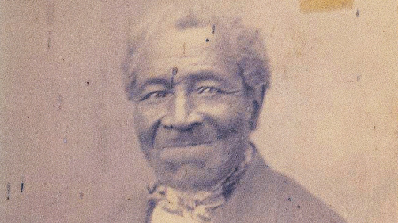 A weathered 1800s portrait photo shows a grey haired man in a jacket and bow tie.