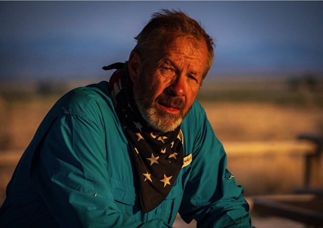 A bearded man wearing a teal shirt and black star-patterned bandana around his neck sits outdoors in warm evening light.