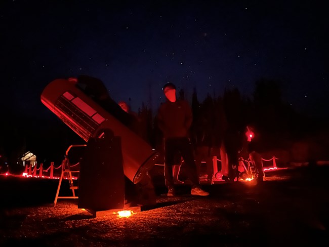 A man stands next to a telescope illuminated by red light before a starry sky.