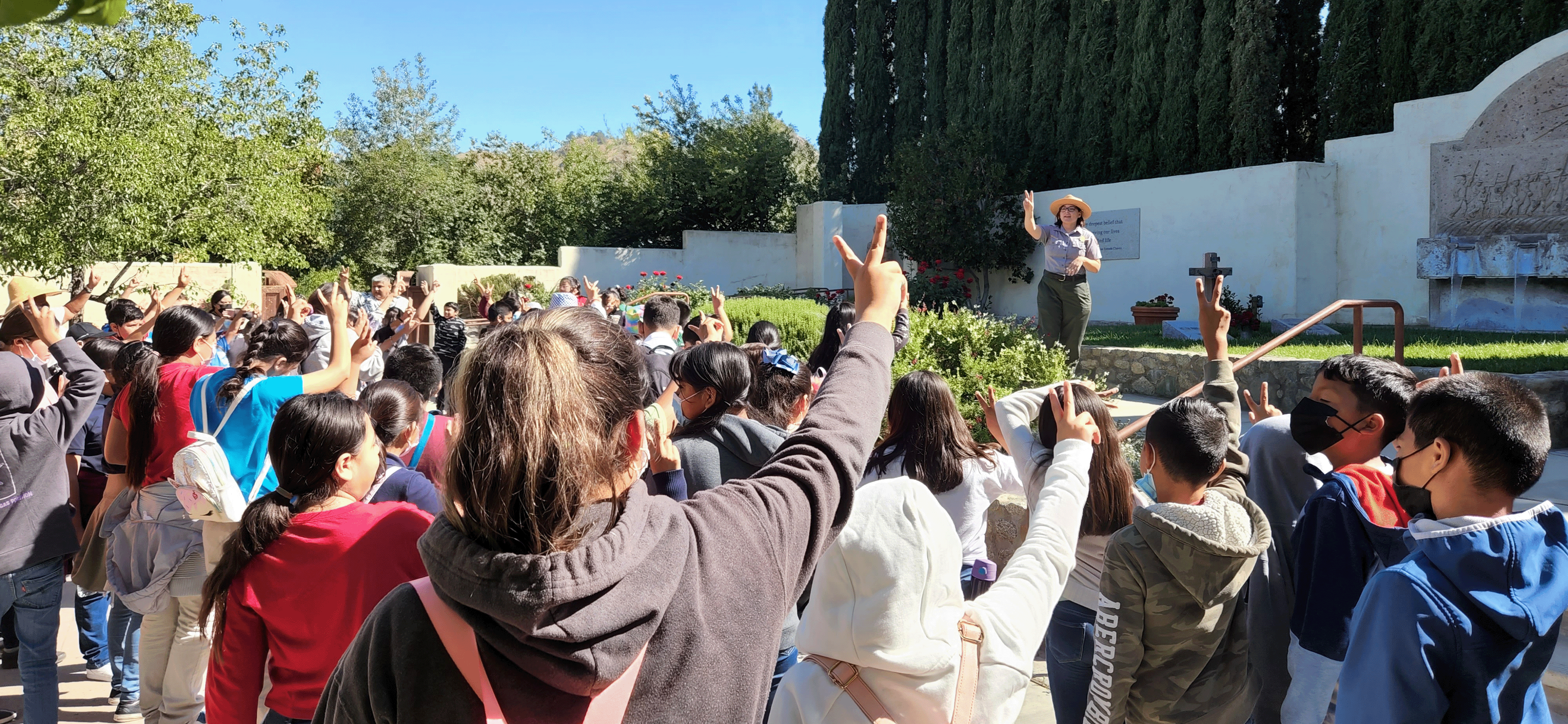 A park ranger stands in front of a group of school children in an outdoor garden holding two fingers in the air while the children do the same