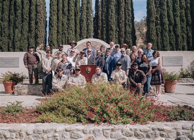 A large group of people stand on either side of a podium, with some uniformed people kneeling in front of it, with a concrete wall and tall cypress trees in the background.