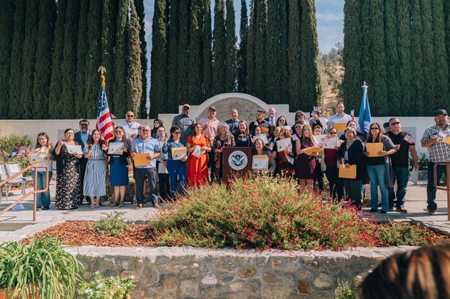 A large group of people standing around a podium with a wall and tall cypress trees in the background.