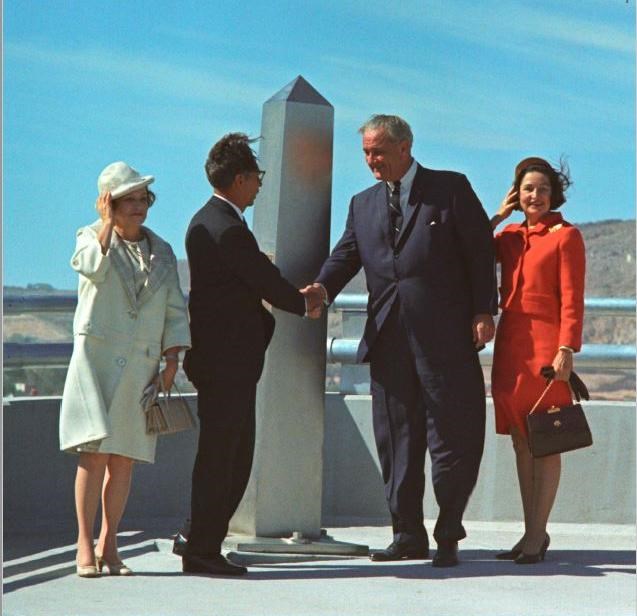 Presidents Díaz Ordaz and Johnson shake hands in front of a grey boundary monument in this color photo. Their wives stabd on either side of them, holding their hats against the breeze.