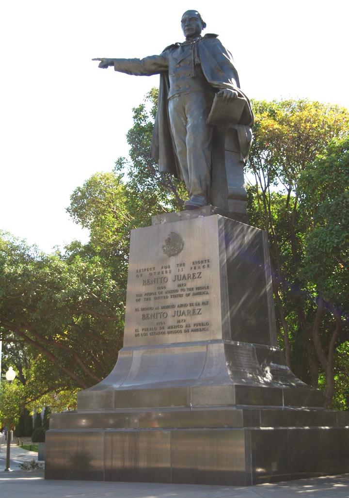 Una estatua en bronce de un hombre llevando un manto y cargando un libro en la mano izquierda y señalando con la mano derecha. La estatua está instalada en un pedestal grande de metal inscrito.
