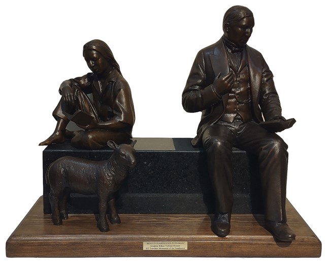 Small bronze maquette showing a youth seated on the left side of a bench with a sheep at his feet and a well dressed adult man to the right. Both are reading books.