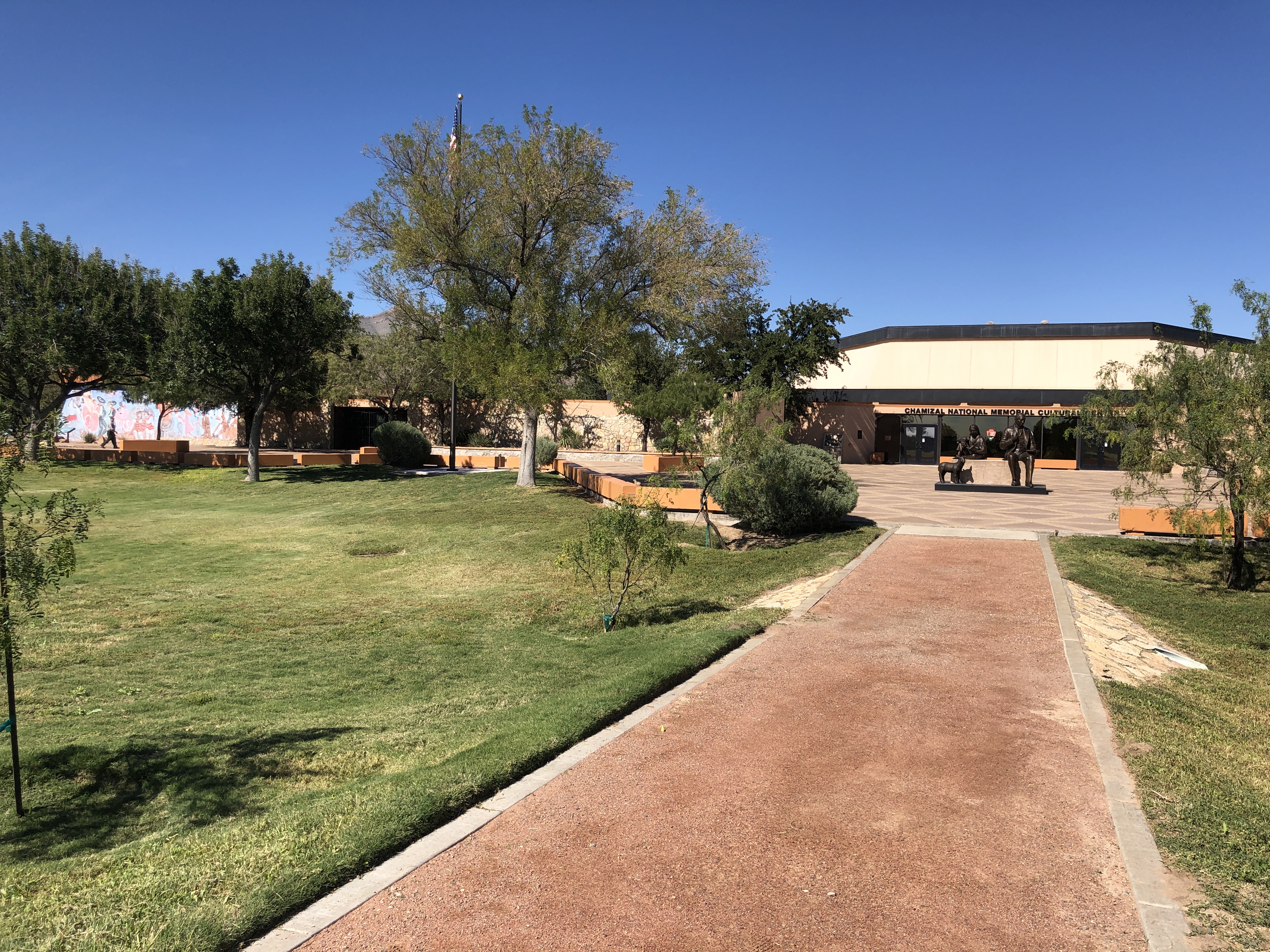 A red gravel path crosses a stretch of green lawn to join an open paved area in front of a sand-colored building with a bright mural on its far left side. On the esplanade in front of the building is a bronze sculpture of two figures seated on a bench.