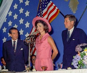 Jacqueline Kennedy stands behind a microphone, flanked by JFK and President Lopez Mateos