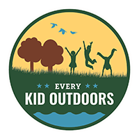 Round logo with a green field with two brown trees. Blue water feature in the foreground, three kids jumping in the field and three birds in flight.