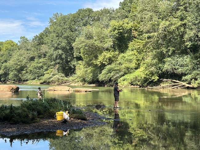 Two people fishing in shallows of river.