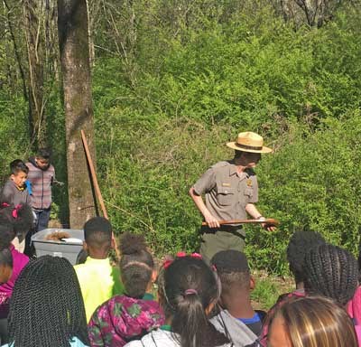 A park ranger talks to a group of students in front of green bushes