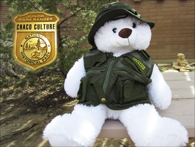 A white stuffed animal bear wearing a Junior Ranger vest and hat. An image of a Junior Ranger badge is next to it.
