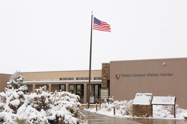 A tan building identified as Chaco Canyon Visitor Center in text, with snow covered plants and a flag flying in the foreground.