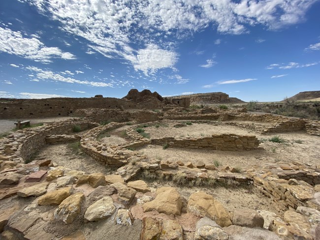 Remains of three concentric circular masonry walls with the desert mesa and white clouds in the background.