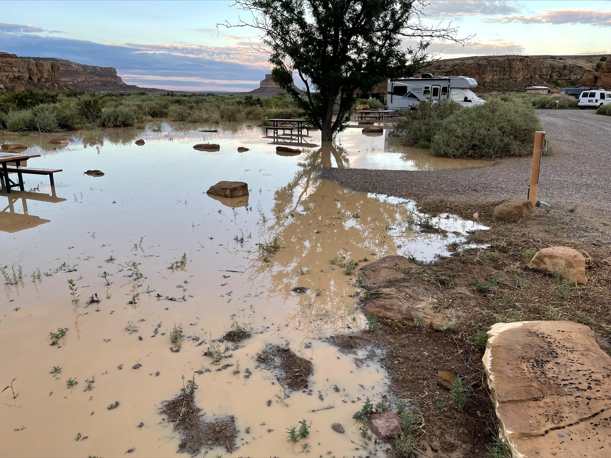 A huge muddy collection of water floods a campground, with an RV and cars in other campsites, and a picnic table partially under the water.
