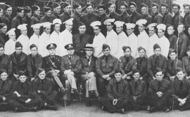 Group photograph of men in dark colored uniforms with several men in white kitchen uniforms in the center row