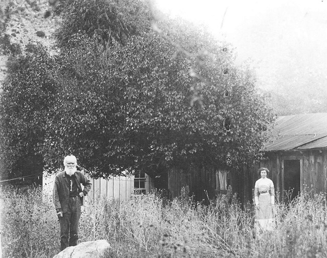 Black and white photo of an older man with a white beard and a young woman standing in front of a log cabin.