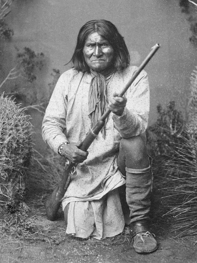Black and white portrait of man with shoulder-length hair, kneeling, holding a rifle, and staring directly at the camera.