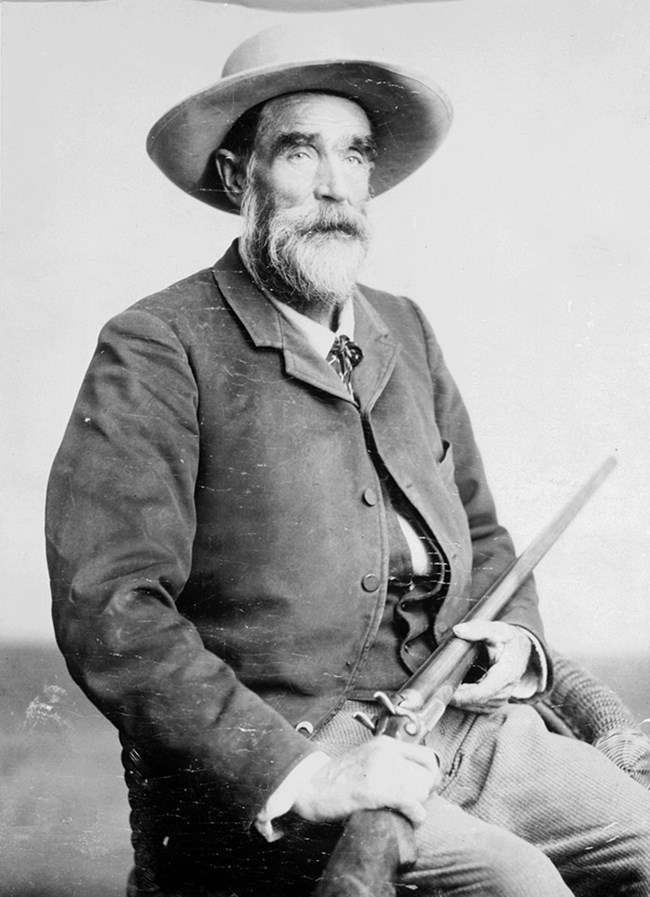 Black and white portrait of a seated man, holding a rifle. The man is wearing a hat and has a white beard.