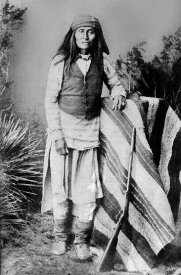 Black and white photograph of a long-haired man wearing pale clothes, a dark vest, and leaning against a table covered in a striped blanket, with a rifle leaning against the blanket.