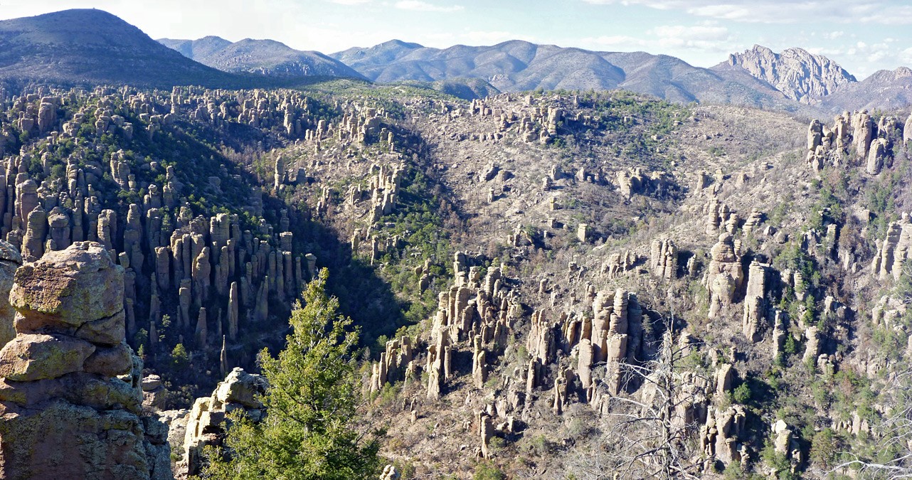 Rocky mountain behind rock pinnacles and spires, interspersed with trees.
