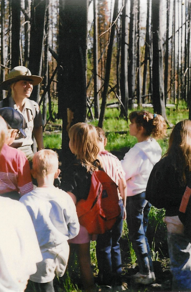 A park ranger stands in front of a standing group of children in a stand of charred trees.