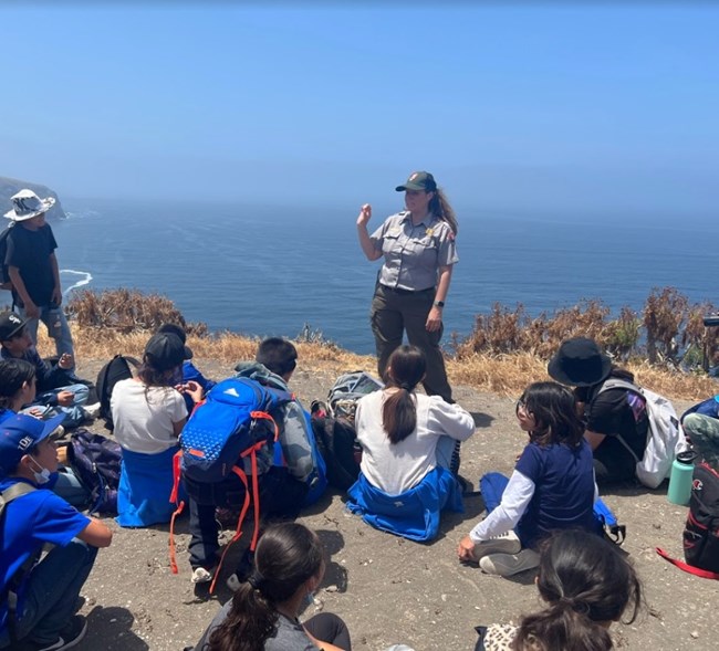 A park ranger stands in front of a group of children sitting on the ground. Behind the ranger are some plants and the ocean