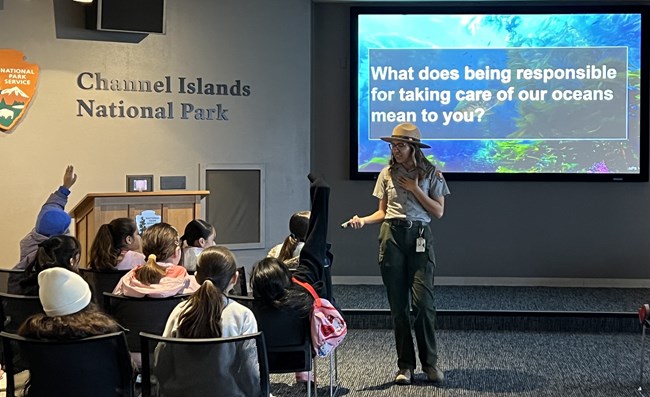 Park Ranger stands in front of a group of seated students with a screen behind that reads "What does being responsible for taking care of our oceans mean to you?"