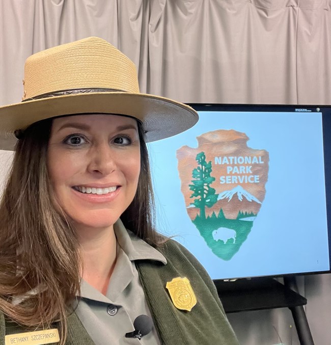 a female ranger with a flat hat on smiles in front of a screen with the NPS arrowhead on it.