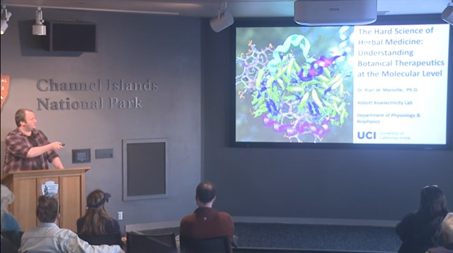 man at podium talking and looking at screen with plant image