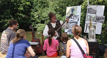 A man in 19th century civilian clothing plays a banjo for a family.