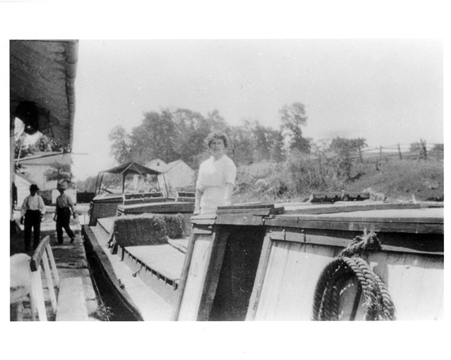 Historic Photo of a woman dtanding on a canal baot at four locks, with buildings and people walking in the background.