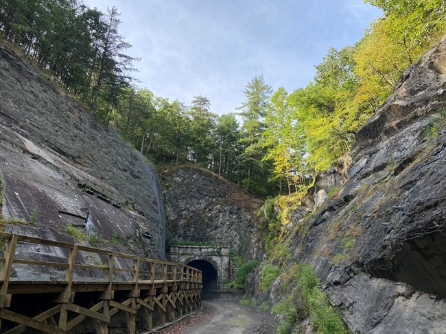 A wooden boardwalk leads through a ravine with towering rock on either side and into a historic stone tunnel