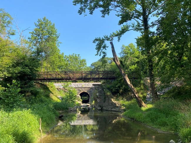 A metal bridge spans the ramains of a historic stone culvert with a stream running below