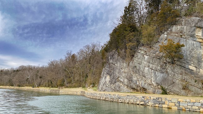 A river flows beside a stone retaining wall holding up a dirt pathway