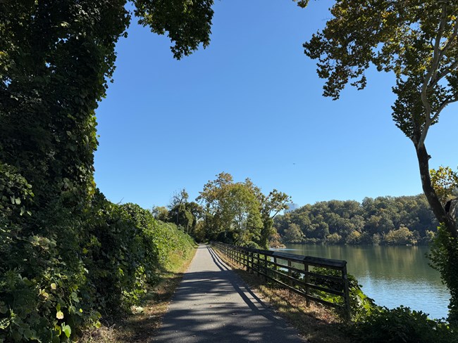 A paved trail with wooden railing leads away from the camera, a river to the right of it and a row of green vegeation to the left.