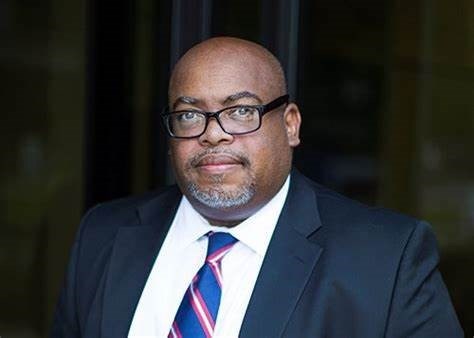 “Portrait of Dr. Brian Mitchell, wearing a dark suit, white shirt, and a red and blue striped tie. He has glasses, a short beard, and a serious expression, standing in front of a blurred background.