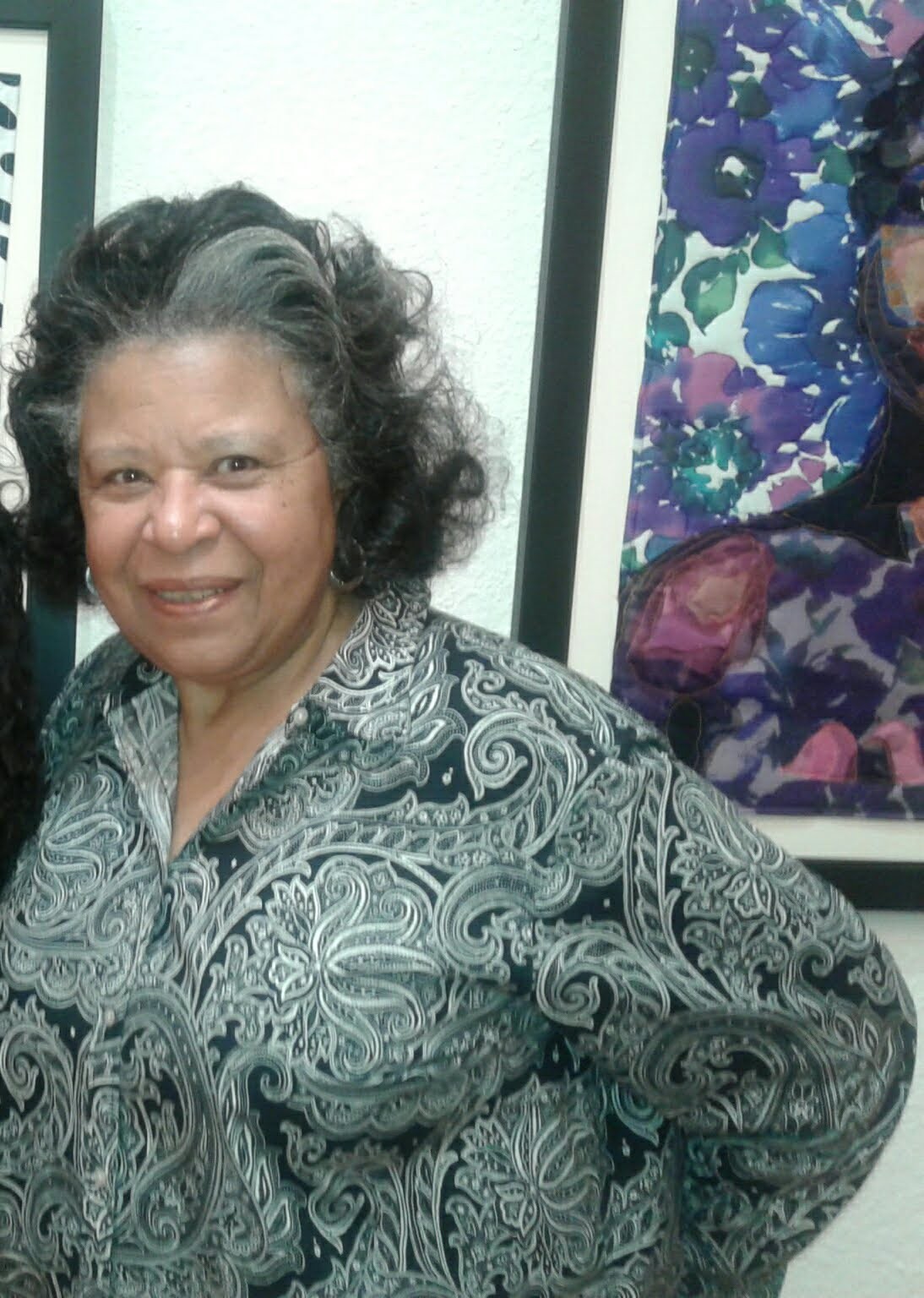Smiling woman with short, curly gray hair wearing a patterned blouse, standing in front of colorful artwork and a white wall.