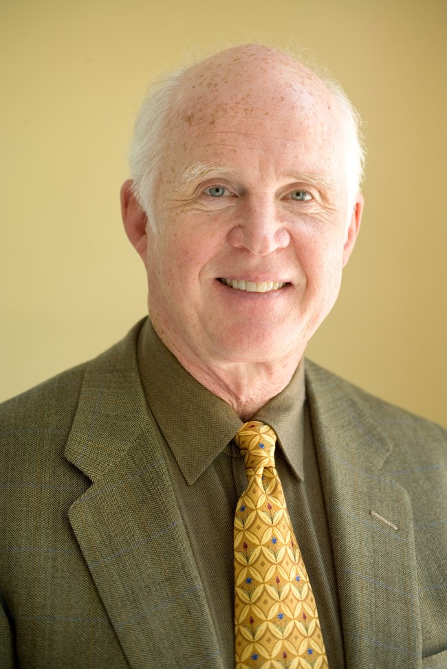 A professional portrait of a man with fair skin and light freckles. He is smiling warmly and wearing a green herringbone suit, a darker green shirt, and a yellow tie with a geometric pattern featuring blue and red accents.