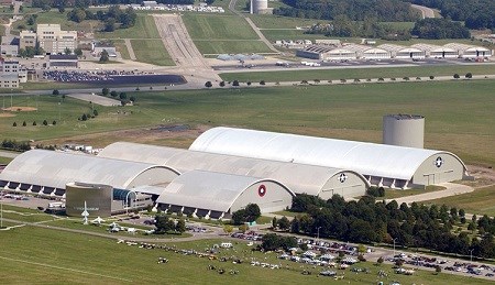 Three large buildings with concave roofs surrounded by grassy fields, a runway in the background and a parking lot in the foreground.