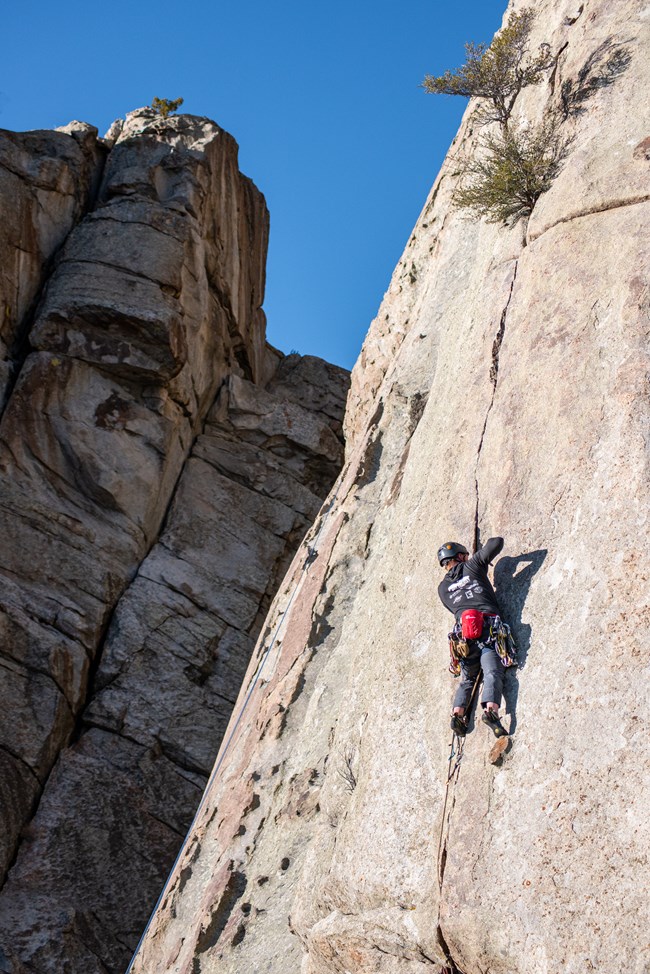 A climber ascends a thin vertical crack, he stands on a small nub while placing protective gear.