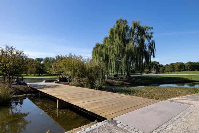 A boardwalk with no railing extends to an island where two visitors sit near the Signers of the Declaration memorial.