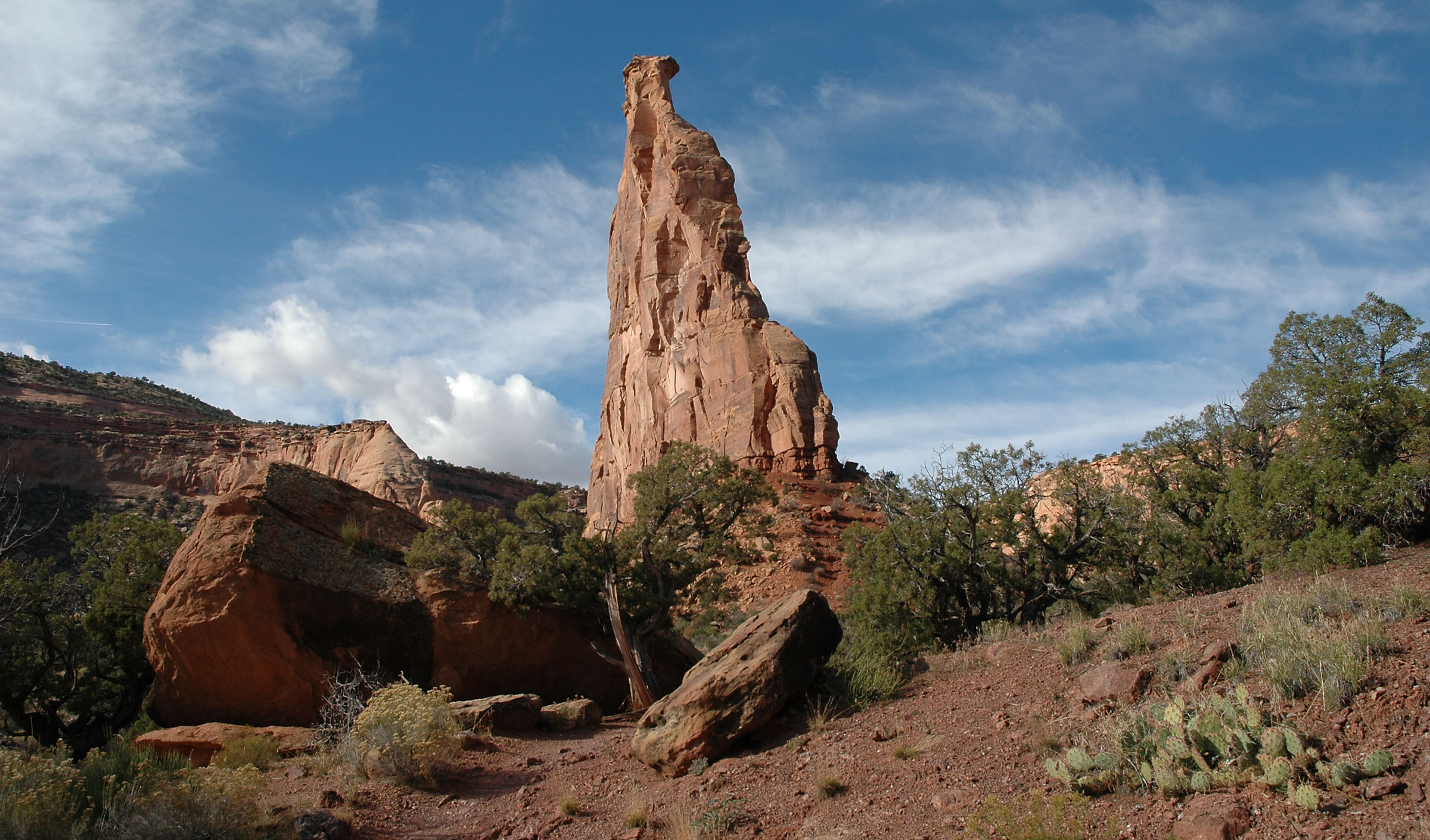 Independence Monument stands out against the sky as the viewer looks up at it from near its base. There are rocks and foliage in the foreground.