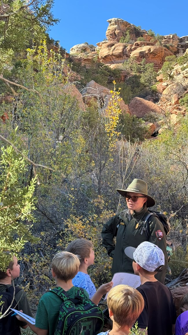 Uniformed park ranger talks to a group of students beneath a rocky cliff and trees.