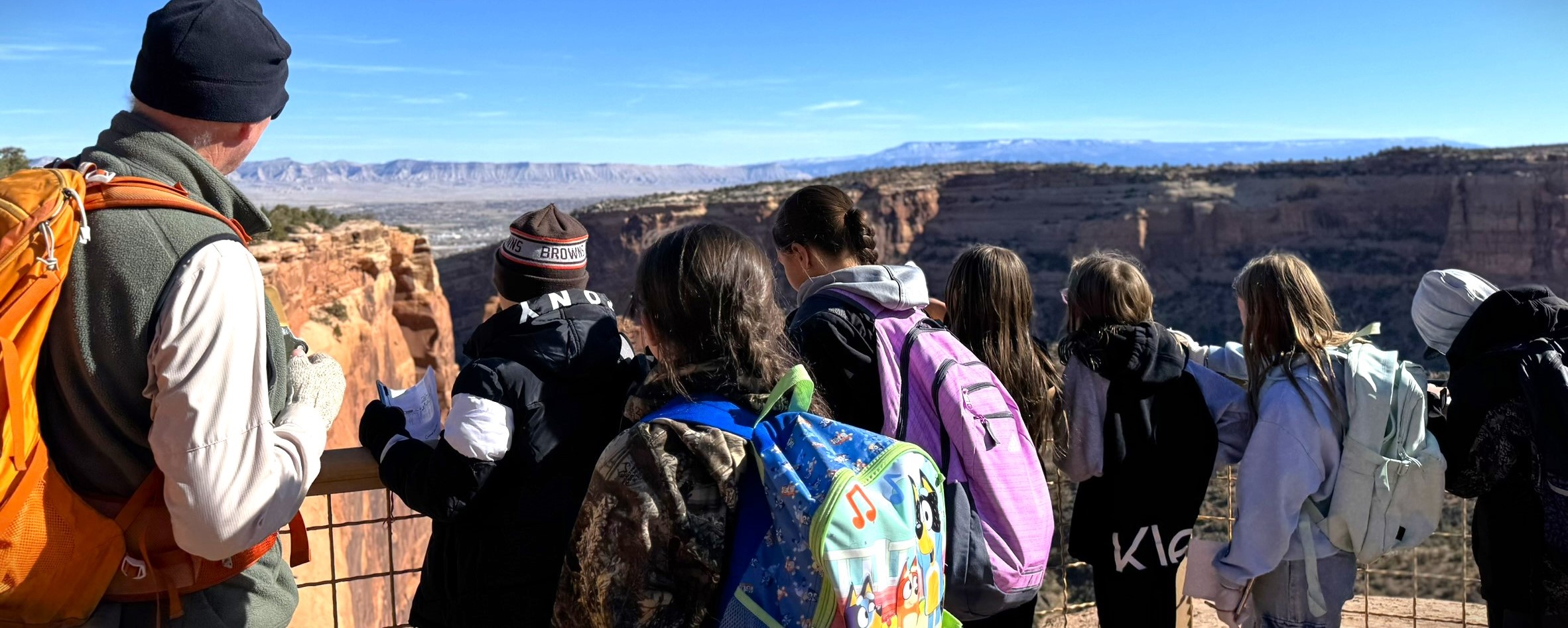A ranger looks away towards desert canyons and valleys, standing next to seven students wearing backpacks.