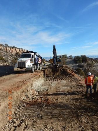 Excavator removes four feet of road base. A dump truck waits along side for the dirt and rock. Two people in safety vests stand in area previously excavated in the foreground of the photo.