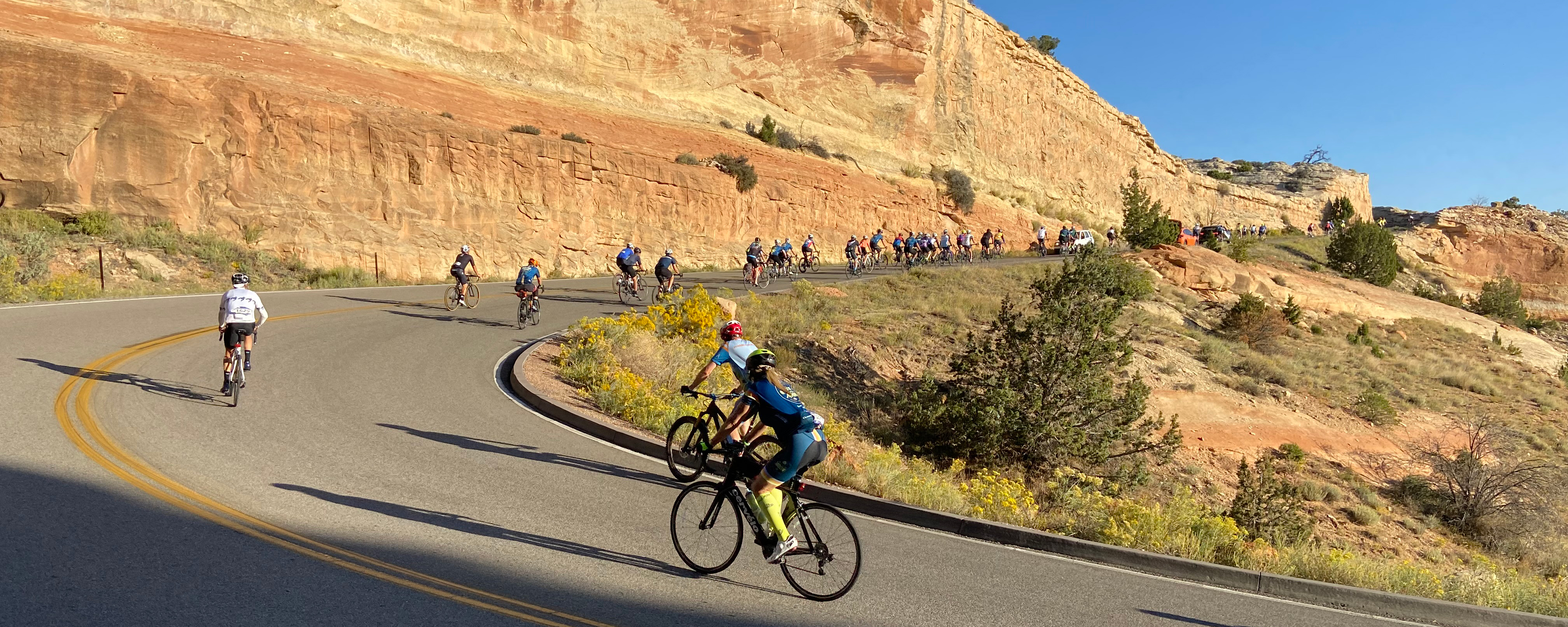 cyclists move uphill on road along orange sandstone cliff