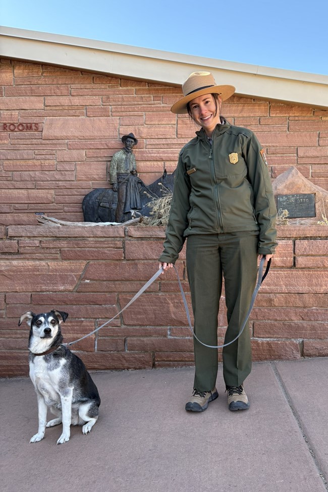 A ranger in uniform holds a leash attached to a small dark dog on pavement.