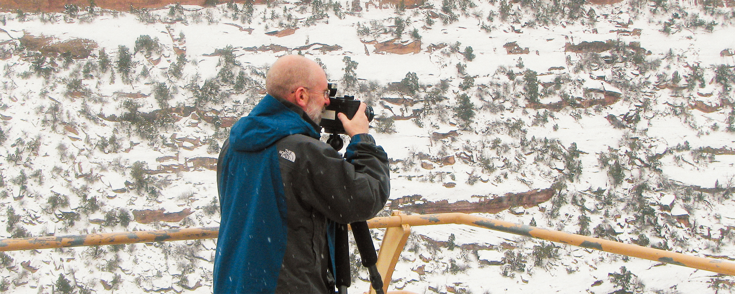 bald man in winter coat holds camera pointed towards snowy canyons
