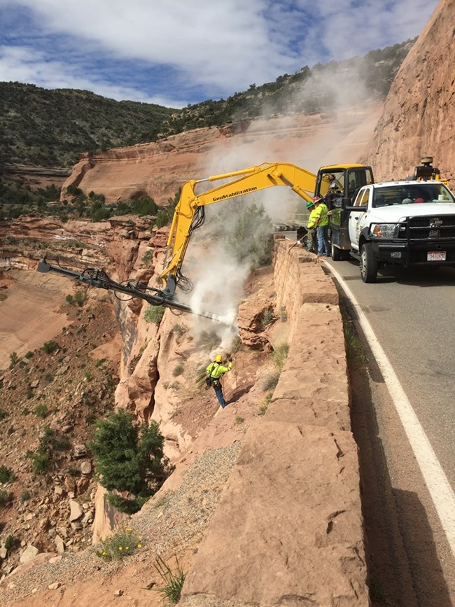 Heavy equipment extends drill over edge of road to repair culvert. Worker on ropes below road to direct work.