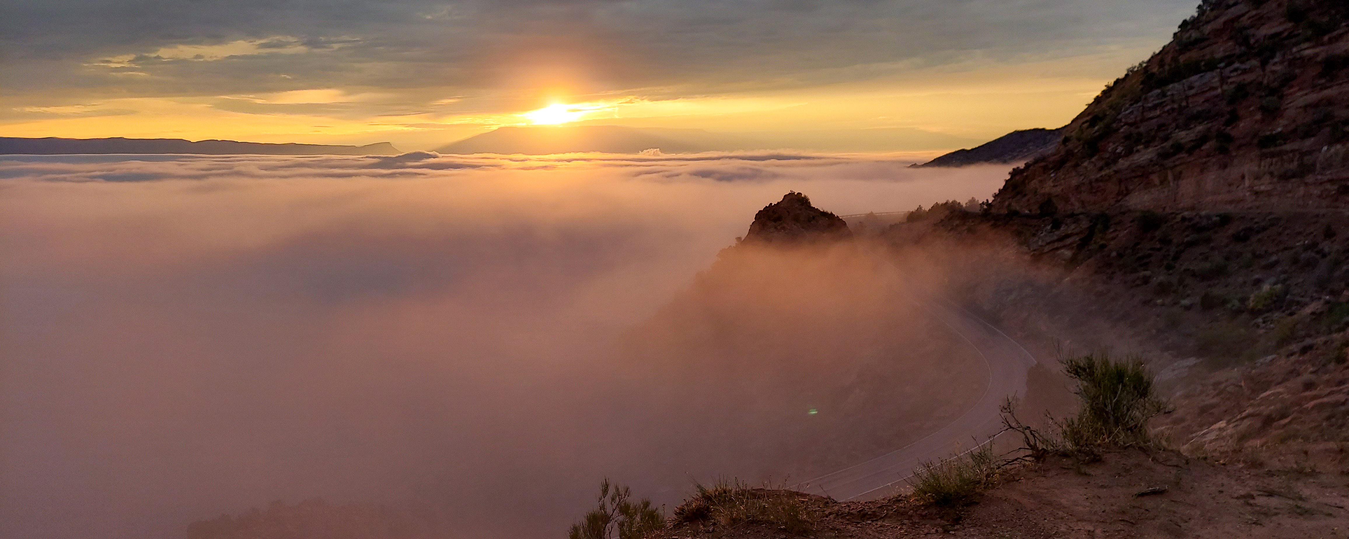 clouds below hide canyon bottoms as sun rises above mesa in distance